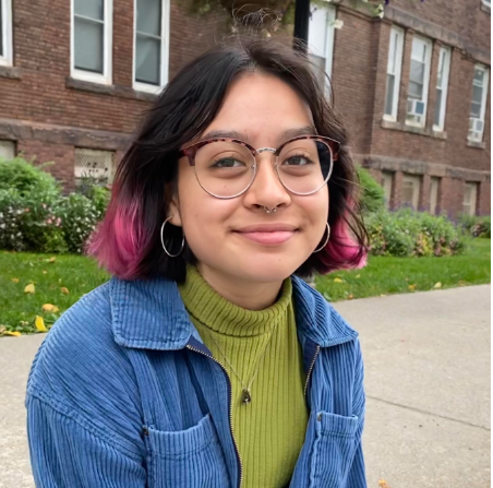 Person with glasses and dark hair with dyed pink ends seated with a brick building in the background