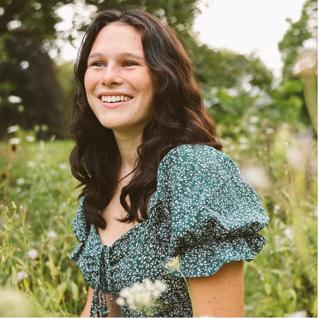 Photo of person with long dark hair, looking away from camera, standing in a garden