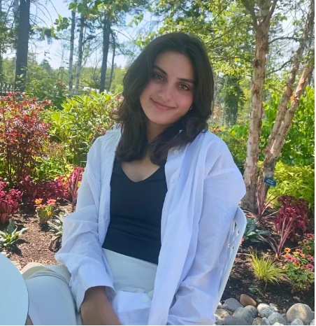 Photo of person with long dark hair, smiling, seated in a sunny garden