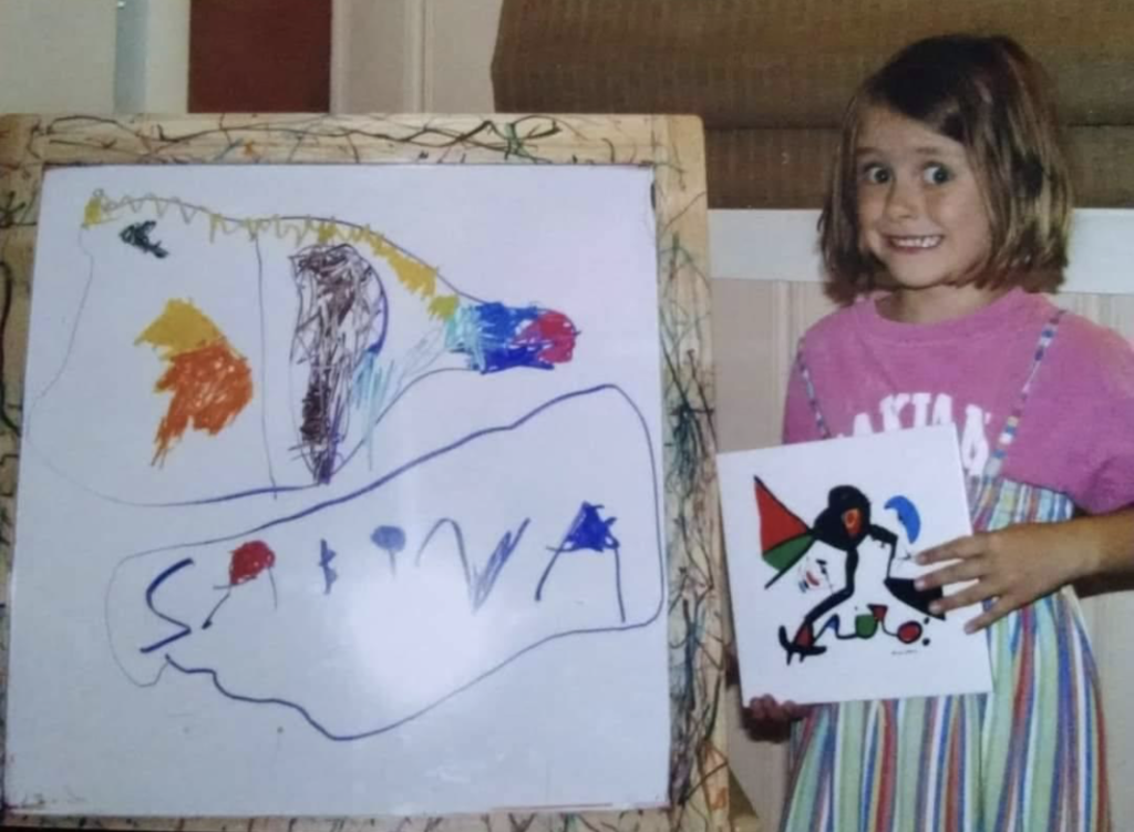 A child holding a ceramic painted hot plate next to their recreation of the design on an easel. They are wearing a pink T-shirt under a striped green dress. They have shoulder-length hair and an awkward four-year-old smile.