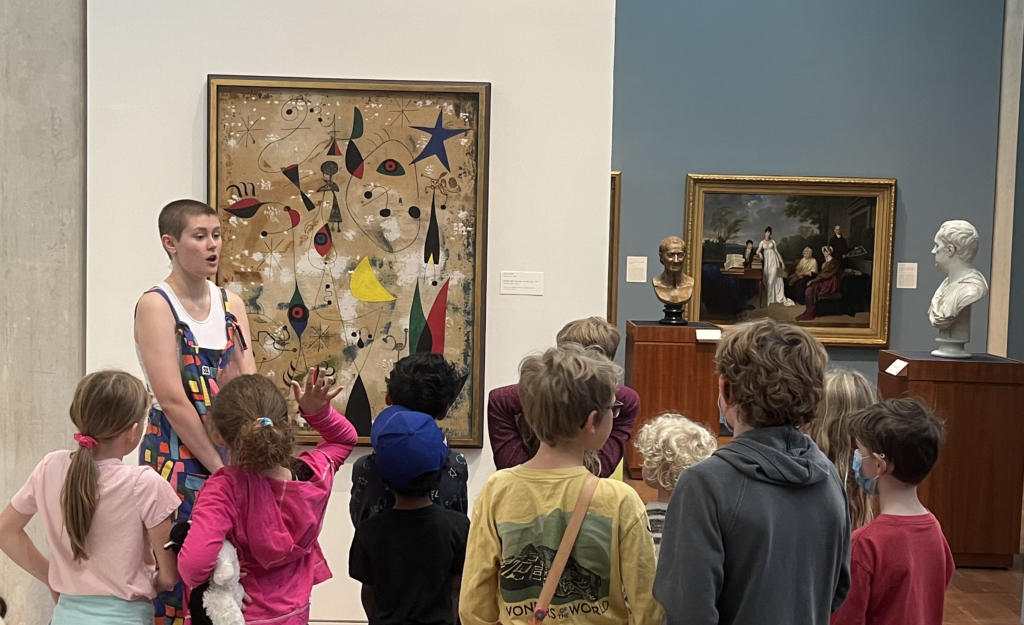 Photo of a college student standing in an art gallery, talking to a group of young children.